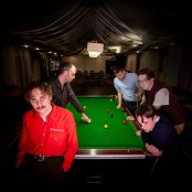 The five members of the band Hotel Lux stood around a pool table in a dimly lit room